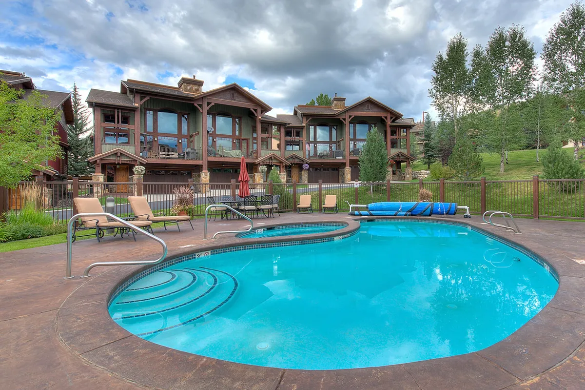 A view of the community's outdoor heated pool surrounded by lounge chairs and trees, with the townhome's building facade visible. The setting is leisurely and designed for socializing or relaxing in both summer and winter.