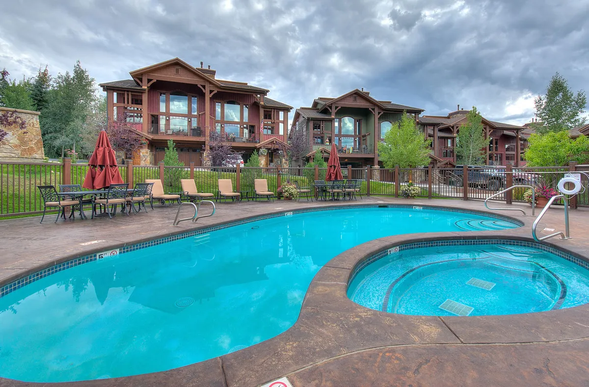 This image showcases an inviting outdoor heated swimming pool and an adjacent hot tub, surrounded by lush greenery and lounge chairs. The resort buildings are visible in the background, creating a sense of community and luxury. The tranquil mood is enhanced by the soft outdoor lighting, and the setting is ideal for relaxing, swimming, or enjoying poolside gatherings.