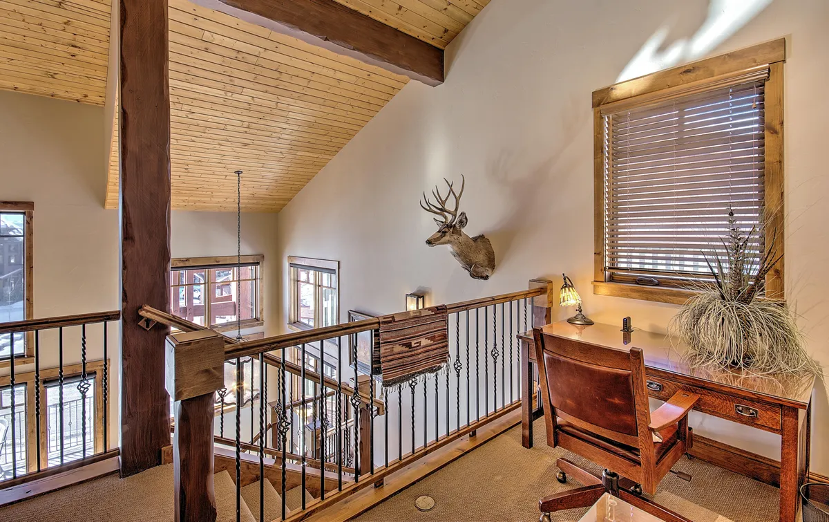 This image captures a cozy, rustic loft space featuring a wooden railing, a work desk with a chair, a mounted deer head, and large windows that bring in natural light and offer inspiring alpine views. The setting is perfect for someone working remotely or seeking a quiet retreat.