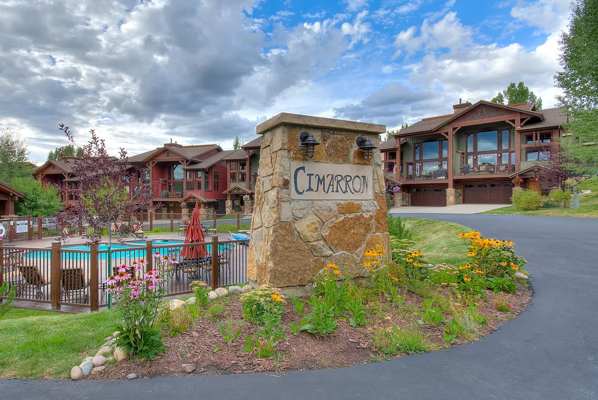 This exterior image captures the Cimarron property sign, a stone structure, the inviting swimming pool surrounded by lush flowers, and the brown buildings that comprise the community. The scene exudes a welcoming, resort-style ambiance perfect for leisure, with visible spaces for sunbathing and gathering.