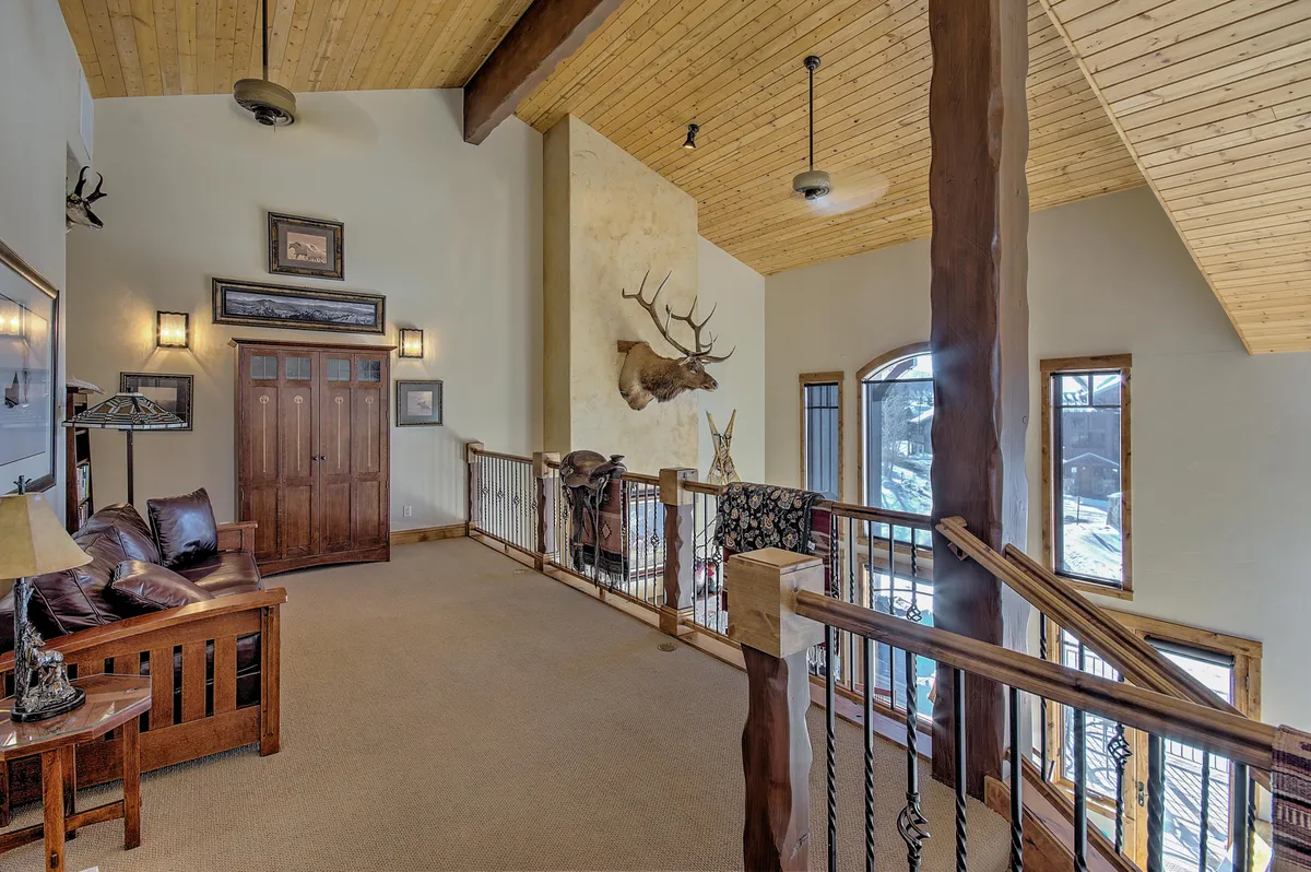 The loft area features a comfortable leather sofa, wooden railing, a mounted deer head, and expansive windows that flood the space with light. The setting encourages relaxation, socializing, or unwinding with a good book or movie.