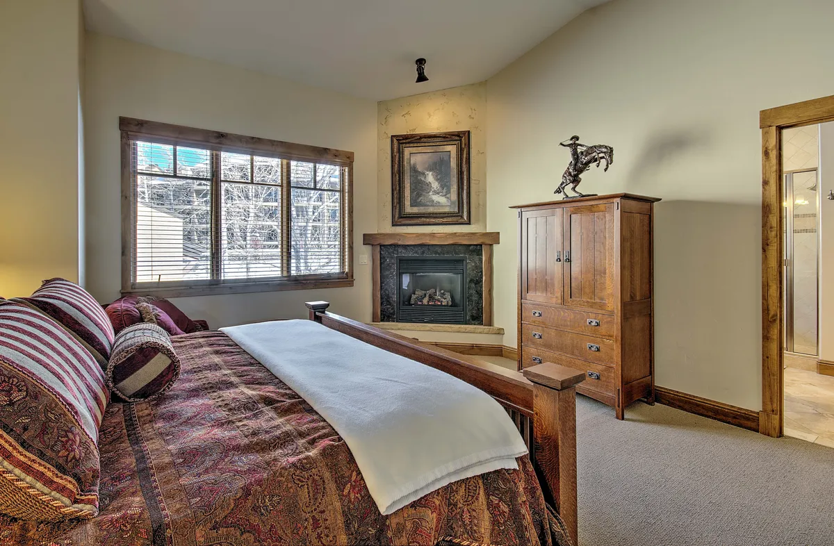 A spacious bedroom featuring a bed with decorative bedding, a cozy fireplace, a large window allowing in natural light, and wooden furniture. The room exudes tranquility and comfort, ideal for restful nights or relaxing by the fire.