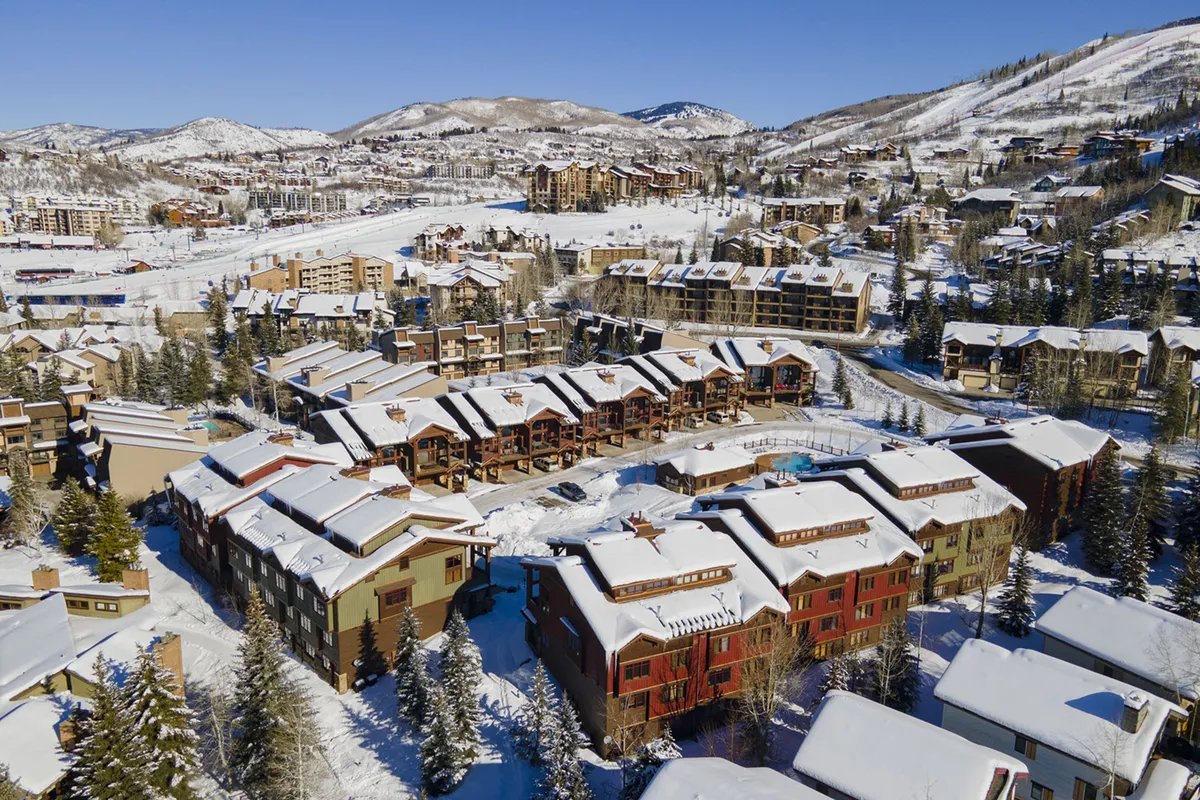 An exterior shot of snow-covered rooftops, pine trees, mountain backdrop, and residential buildings. The image evokes Steamboat's winter charm and proximity to mountain activities like skiing and snowboarding.