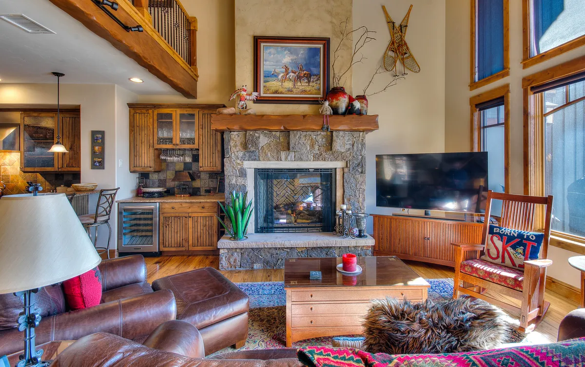 The image showcases the inviting living area of the townhome, featuring a cozy fireplace framed by elegant wooden cabinetry, a comfortable rocking chair, large windows letting in natural light, and tasteful artwork above the mantel. The warm ambiance suggests a perfect spot for relaxing after a day exploring Steamboat Springs.