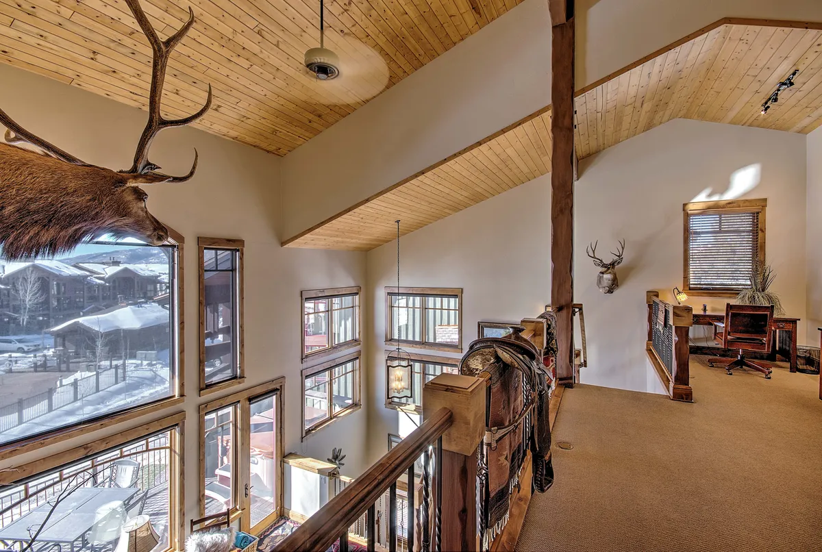 This inviting loft space is characterized by large windows with mountain views, a rustic wooden railing, mounted elk head, and an interior desk setup. The decor is warm and welcoming, blending productivity with leisure.